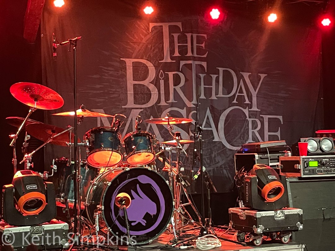 stage with instruments, drums, guitars, lights, sound board, hanging tapestry that says The Birthday Massacre